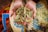A pair of hands, slightly dirtied, cradles a collection of grains or seeds. The background features a vibrant woven basket with a variety of colors including blue, red, yellow, and green, filled with similar grains.