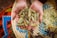 A pair of hands, slightly dirtied, cradles a collection of grains or seeds. The background features a vibrant woven basket with a variety of colors including blue, red, yellow, and green, filled with similar grains.