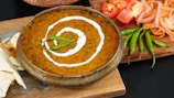A bowl of creamy lentil curry with a swirl of cream on top, garnished with green chilies. It is surrounded by sliced tomatoes, onions, and fresh green chilies on a wooden board, accompanied by pieces of naan bread.
