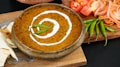 A bowl of creamy lentil curry with a swirl of cream on top, garnished with green chilies. It is surrounded by sliced tomatoes, onions, and fresh green chilies on a wooden board, accompanied by pieces of naan bread.