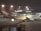 A large passenger airplane with the logo of Qantas on the side is parked on the tarmac at an airport. The scene is set during the night with artificial lights illuminating the aircraft and the terminal building in the background. The airport terminal has large glass windows and is bustling with activity.