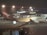 A large passenger airplane with the logo of Qantas on the side is parked on the tarmac at an airport. The scene is set during the night with artificial lights illuminating the aircraft and the terminal building in the background. The airport terminal has large glass windows and is bustling with activity.