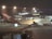 A large passenger airplane with the logo of Qantas on the side is parked on the tarmac at an airport. The scene is set during the night with artificial lights illuminating the aircraft and the terminal building in the background. The airport terminal has large glass windows and is bustling with activity.