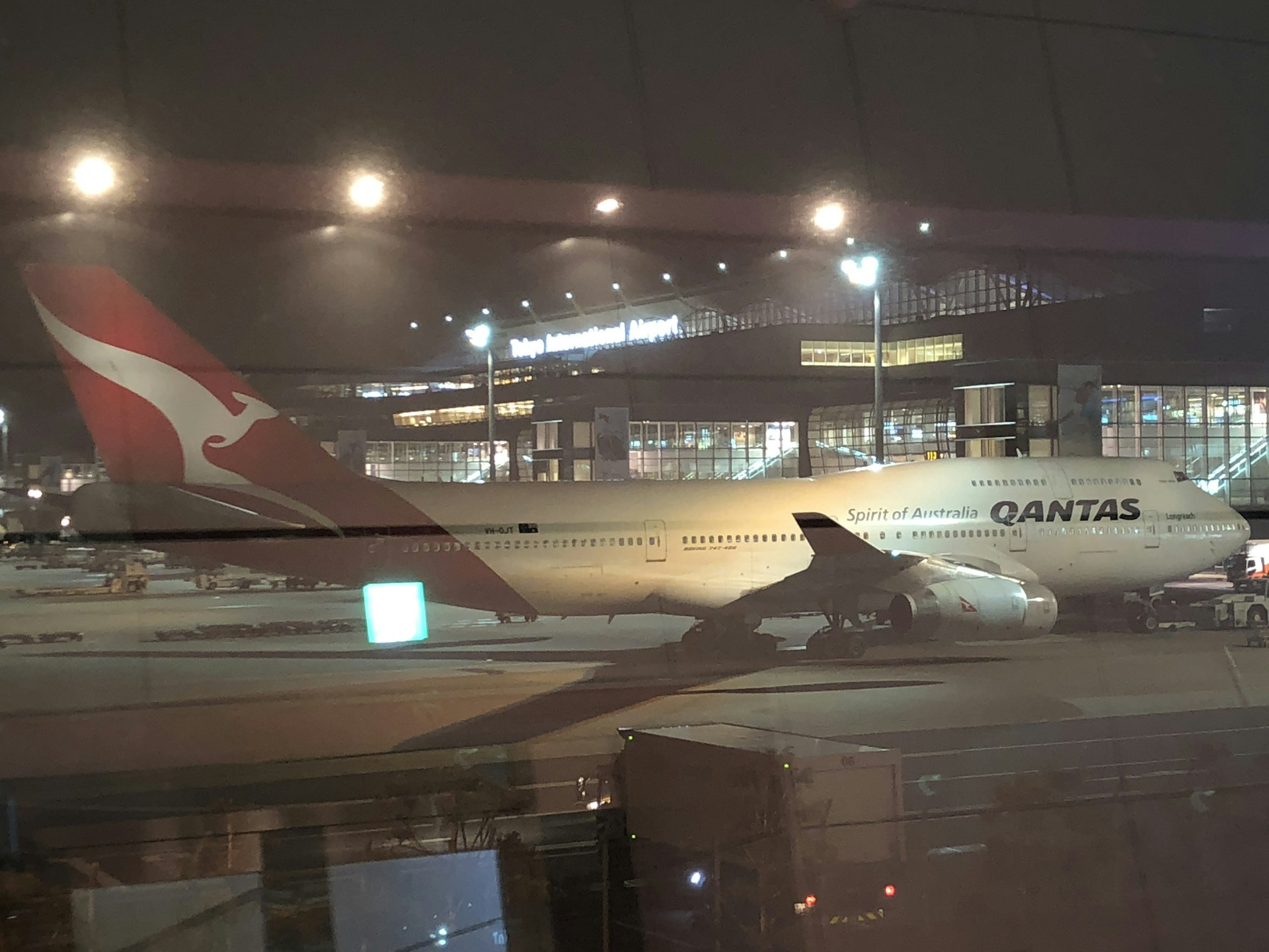 A large passenger airplane with the logo of Qantas on the side is parked on the tarmac at an airport. The scene is set during the night with artificial lights illuminating the aircraft and the terminal building in the background. The airport terminal has large glass windows and is bustling with activity.