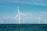 Several large wind turbines are installed in the ocean, with clear blue skies above and the deep blue ocean below. The turbines are evenly spaced across the horizon, suggesting a wind farm dedicated to renewable energy generation.