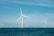 Several large wind turbines are installed in the ocean, with clear blue skies above and the deep blue ocean below. The turbines are evenly spaced across the horizon, suggesting a wind farm dedicated to renewable energy generation.