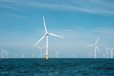 Several large wind turbines are installed in the ocean, with clear blue skies above and the deep blue ocean below. The turbines are evenly spaced across the horizon, suggesting a wind farm dedicated to renewable energy generation.