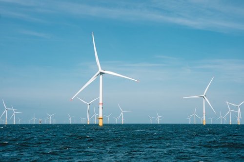Several large wind turbines are installed in the ocean, with clear blue skies above and the deep blue ocean below. The turbines are evenly spaced across the horizon, suggesting a wind farm dedicated to renewable energy generation.