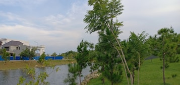 A serene landscape featuring a calm body of water bordered by green grass and trees. A modern house is visible in the background, along with a blue fence lining the property. The sky is partly cloudy with hints of blue and white, contributing to a tranquil atmosphere.