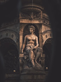 A classical stone statue of a seated person is prominently displayed in a decorative architectural niche. The statue is detailed, showcasing intricate craftsmanship with a sense of realism. The background features ornate carvings and the dark setting highlights the statue with a dramatic lighting effect.