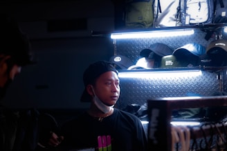 A person wearing a black cap and dark clothing stands in a dimly lit retail environment, with several caps and hats displayed on shelves behind them. The lighting focuses on the middle shelf, highlighting the products while the foreground and background remain darker.