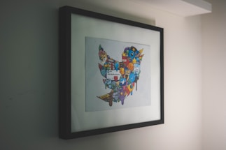 A framed artwork hangs on a wall, featuring a collection of colorful, cartoon-like icons and symbols forming a distinct shape. The artwork is enclosed in a simple black frame, with the primary focus being on the vibrant imagery within.