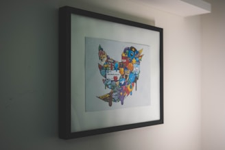 A framed artwork hangs on a wall, featuring a collection of colorful, cartoon-like icons and symbols forming a distinct shape. The artwork is enclosed in a simple black frame, with the primary focus being on the vibrant imagery within.