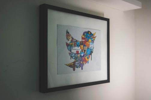 A framed artwork hangs on a wall, featuring a collection of colorful, cartoon-like icons and symbols forming a distinct shape. The artwork is enclosed in a simple black frame, with the primary focus being on the vibrant imagery within.