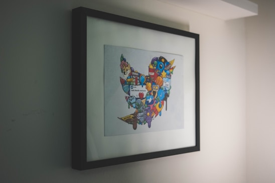 A framed artwork hangs on a wall, featuring a collection of colorful, cartoon-like icons and symbols forming a distinct shape. The artwork is enclosed in a simple black frame, with the primary focus being on the vibrant imagery within.