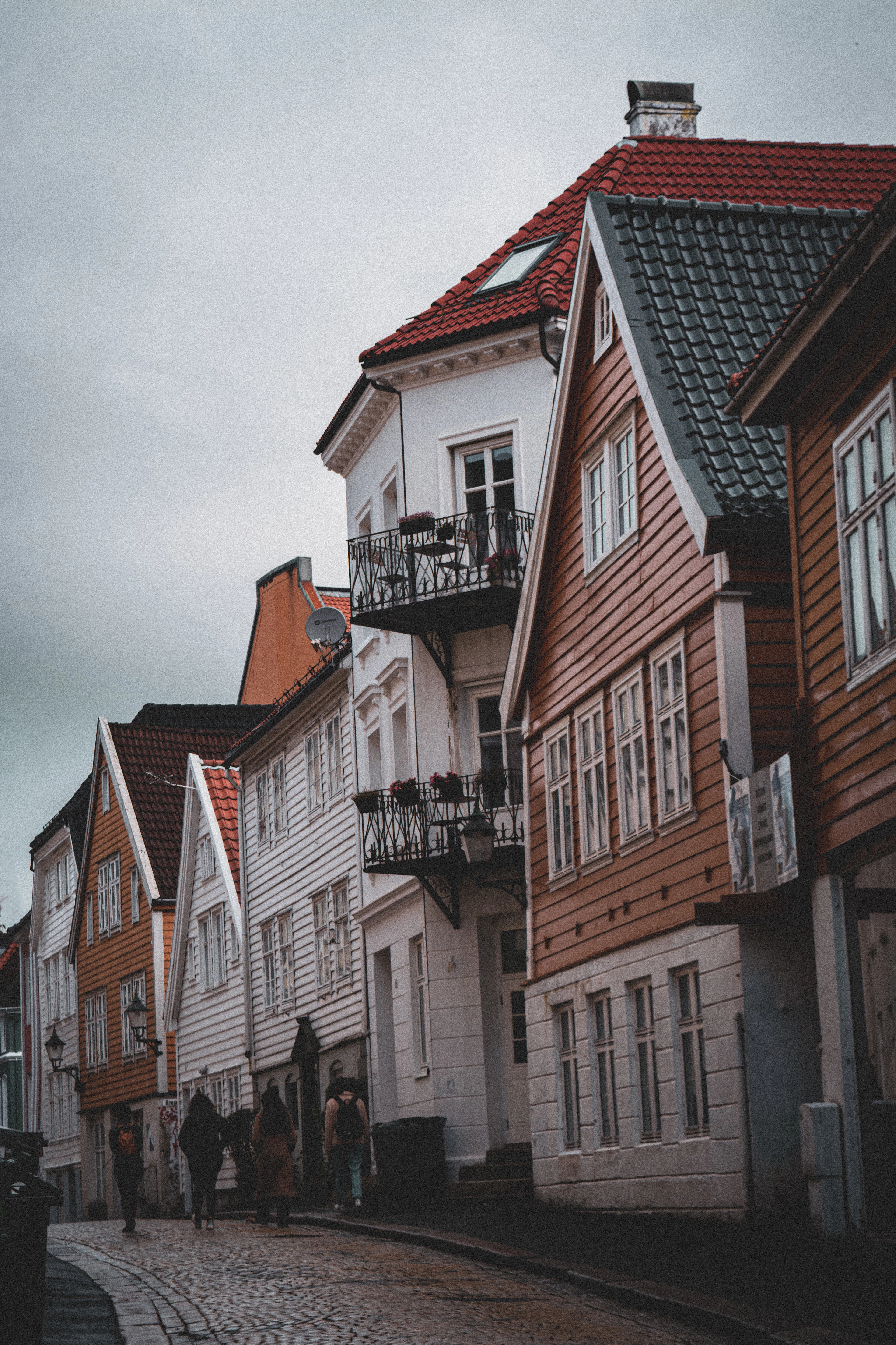 A grey, damp afternoon on a Bergen street, people walking the wet cobbles past tall wooden houses in ochre, rust and white beneath a low pale sky.