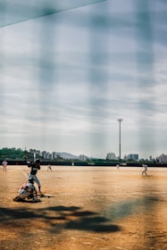 A baseball game in progress on a dirt field with players positioned across the field. The image features a batter preparing to swing at a pitch, with a catcher crouched behind him. The outfield is sparsely populated, and there are tall city buildings visible in the background against a partly cloudy sky.