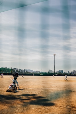 A baseball game in progress on a dirt field with players positioned across the field. The image features a batter preparing to swing at a pitch, with a catcher crouched behind him. The outfield is sparsely populated, and there are tall city buildings visible in the background against a partly cloudy sky.