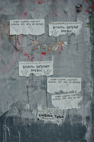 Weathered gray wall with several torn and taped posters containing messages in Russian. Each poster features simple line drawings and handwritten text. The wall itself is cracked and slightly peeling, with traces of paint and texture.