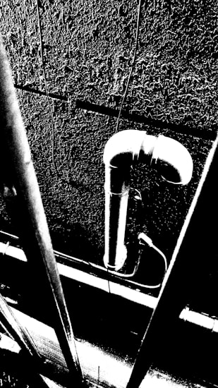 A high-contrast black and white image featuring a vertical pipe on a textured wall. The pipe has a curved joint at the top, with several vertical bars in the foreground. The image conveys a strong sense of structure and industrial design.