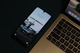 A smartphone is placed beside a laptop on a dark surface. The smartphone's screen displays a wallpaper featuring a car and a jet, while the laptop shows part of a green car image. The laptop's keyboard is visible, featuring typical keys such as 'esc', 'option', and 'command'.