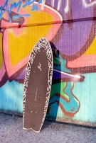 A skateboard with a black grip tape and white patterned edges is propped against a vibrant graffiti-covered wall. The wall features colorful abstract shapes and designs in shades of pink, purple, yellow, blue, and green.