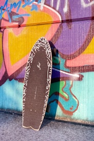 A skateboard with a black grip tape and white patterned edges is propped against a vibrant graffiti-covered wall. The wall features colorful abstract shapes and designs in shades of pink, purple, yellow, blue, and green.