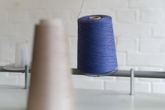 A large spool of blue thread is prominently displayed, hanging on a metal rod. The background shows a blurred brown spool and an out-of-focus brick wall.