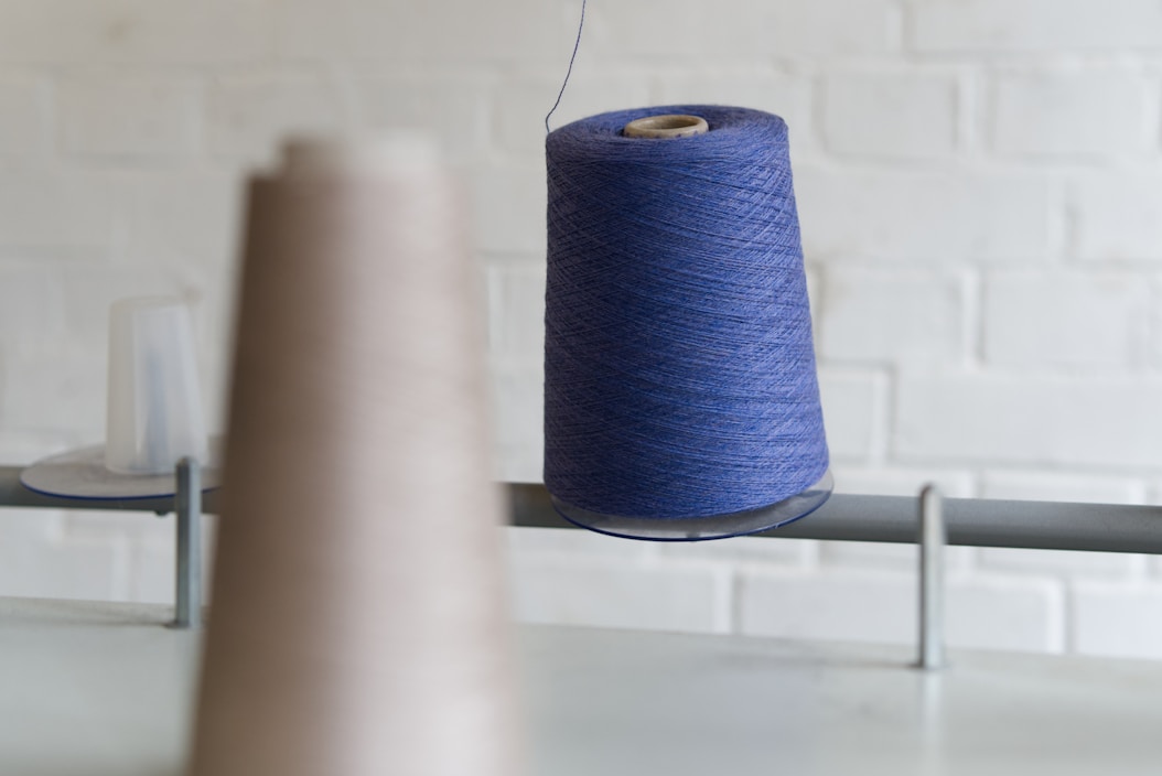 A large spool of blue thread is prominently displayed, hanging on a metal rod. The background shows a blurred brown spool and an out-of-focus brick wall.