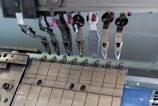 A close-up of a mechanical assembly featuring several metal components and tools, possibly part of a larger industrial or manufacturing machine. Various levers, grips, and fixtures are visible, suggesting a precise and technical function.