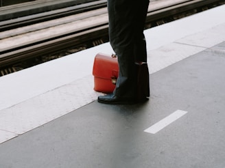 A person stands on a train platform with a bright orange suitcase placed on the ground next to them. The platform has a yellow tactile paving strip for safety.