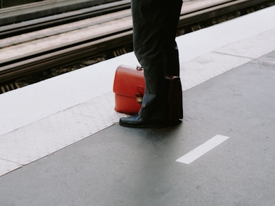 A person stands on a train platform with a bright orange suitcase placed on the ground next to them. The platform has a yellow tactile paving strip for safety.