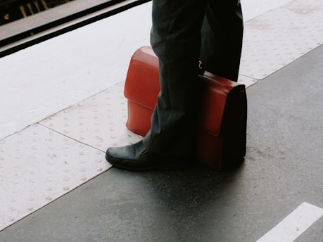 A person wearing black shoes and dark pants is standing on a train platform. Beside them is a brown leather briefcase resting upright.