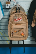 A beige backpack adorned with various patches and logos hangs on a wall. The patches include designs like a flaming brush, a small cartoon character, and the text 'RAW Natural Rolling Papers.' Behind the backpack, a wall with slatted lines and splattered paint serves as the backdrop. Socks with cannabis leaf designs are visible to the left, alongside other hanging apparel.