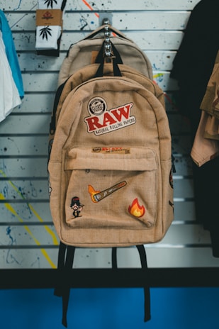 A beige backpack adorned with various patches and logos hangs on a wall. The patches include designs like a flaming brush, a small cartoon character, and the text 'RAW Natural Rolling Papers.' Behind the backpack, a wall with slatted lines and splattered paint serves as the backdrop. Socks with cannabis leaf designs are visible to the left, alongside other hanging apparel.