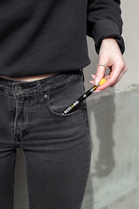 A person wearing a black sweater and dark jeans is holding a disposable vape pen with a visible label. The background appears to be a neutral-colored wall.