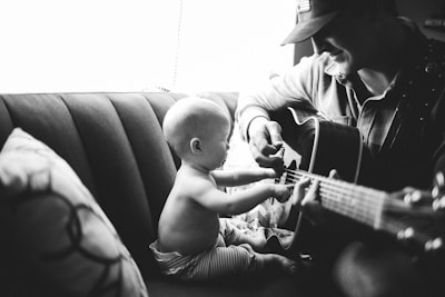 A black and white photograph capturing a tender moment between a baby and an adult. The baby is sitting on a couch, reaching out to touch the strings of a guitar being played by the adult sitting beside them. The adult is wearing a cap and has a gentle smile, likely enjoying the interaction.
