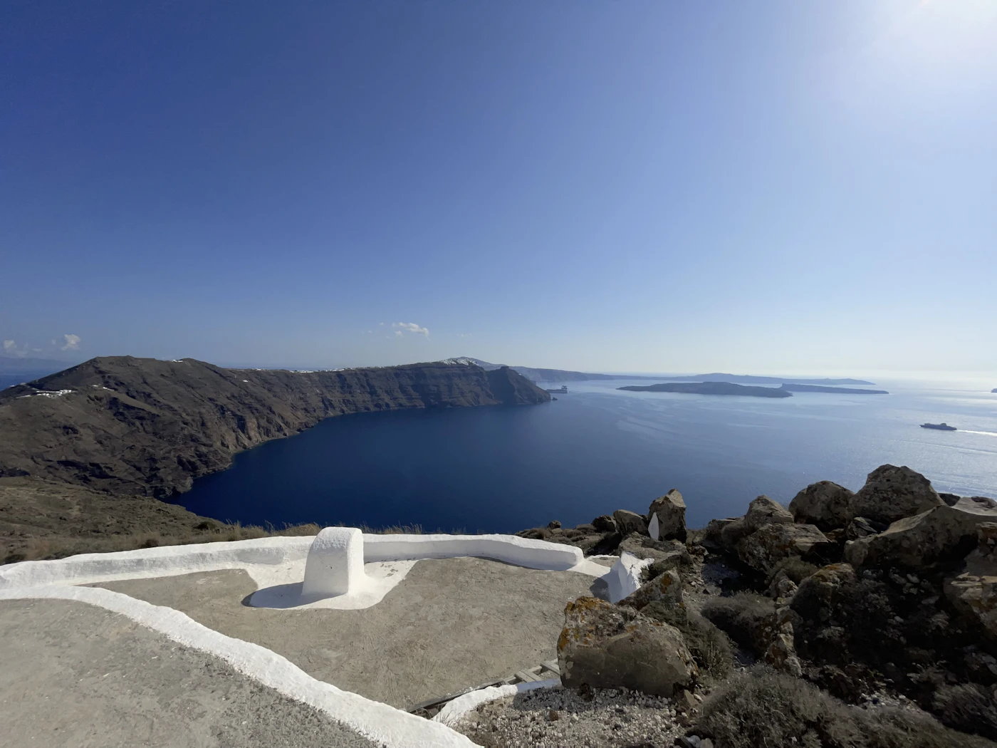 The rim of a volcanic caldera in hard midday sun: a whitewashed parapet in the foreground, black cliffs falling to deep blue water, and hazy islands on the horizon.
