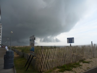 Dark, looming storm clouds hover over a beach boardwalk with tall lamp posts and a wooden fence running parallel to the shore. A few signs are visible along the path. The scene conveys an impending storm approaching the area.