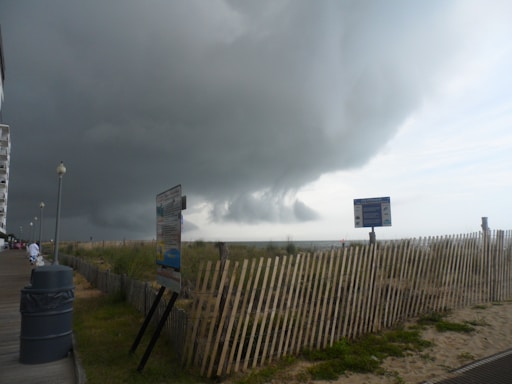 Dark, looming storm clouds hover over a beach boardwalk with tall lamp posts and a wooden fence running parallel to the shore. A few signs are visible along the path. The scene conveys an impending storm approaching the area.