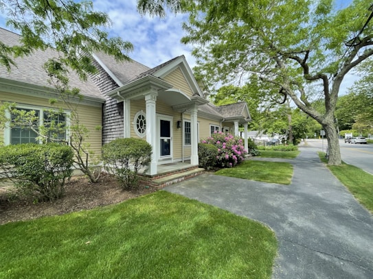 A charming suburban house is surrounded by well-maintained green foliage and a manicured lawn. The house features a gable porch with decorative elements, brick foundation, and light-colored siding. Pink flowering bushes are in bloom near the entrance, framed by tall, leafy trees. A gray asphalt pathway runs alongside the house leading to a quiet street lined with additional trees and greenery.