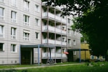 A multi-story residential building with balconies lined with potted plants and a red umbrella. A pathway runs in front of the building, which has grass and trees nearby. Bicycles are parked near the entrance, and the building exterior features a grid-like pattern.