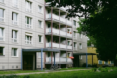 A multi-story residential building with balconies lined with potted plants and a red umbrella. A pathway runs in front of the building, which has grass and trees nearby. Bicycles are parked near the entrance, and the building exterior features a grid-like pattern.