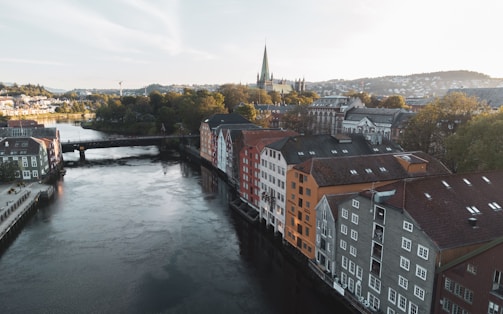 An aerial view of a European cityscape with a river running through, lined with colorful historic buildings with sloped roofs. A bridge spans the river, and a large cathedral with tall spires is visible in the background amidst lush green trees and hills.