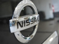 The image features a close-up view of a Nissan car logo, prominently displaying the brand name in chrome lettering on a shiny black surface. The background is blurred out, with some reflections visible on the metal surface.