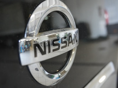 The image features a close-up view of a Nissan car logo, prominently displaying the brand name in chrome lettering on a shiny black surface. The background is blurred out, with some reflections visible on the metal surface.
