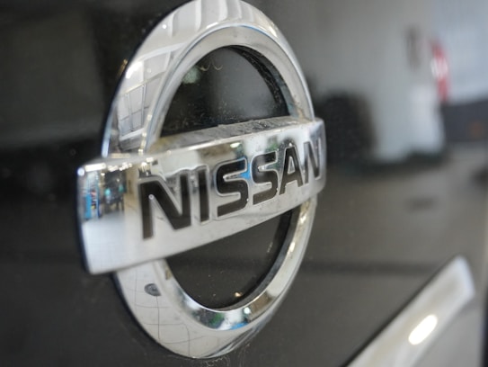 The image features a close-up view of a Nissan car logo, prominently displaying the brand name in chrome lettering on a shiny black surface. The background is blurred out, with some reflections visible on the metal surface.