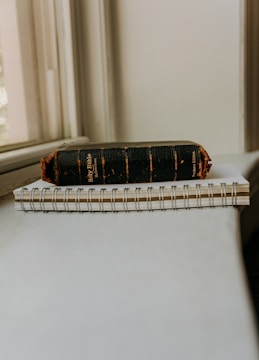 An old, worn-out black Holy Bible with frayed edges rests on top of a spiral-bound notebook. The objects are placed on a light-colored surface near a window, allowing soft natural light to illuminate them.