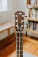 A close-up view of a guitar headstock, featuring tuning pegs and strings. The guitar is placed indoors with a blurred background showing wooden floors and a bookshelf.