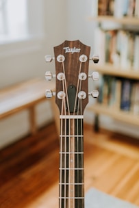A close-up view of a guitar headstock, featuring tuning pegs and strings. The guitar is placed indoors with a blurred background showing wooden floors and a bookshelf.